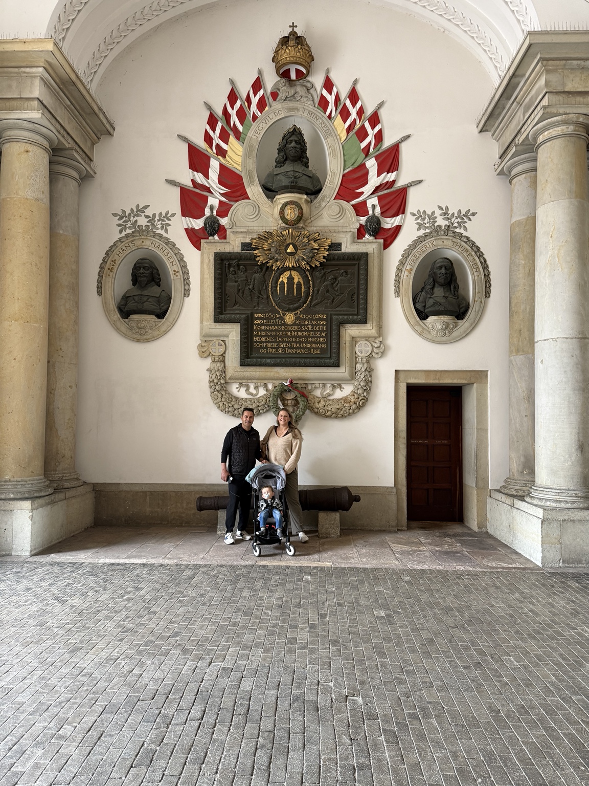 Jeni and Hassan with their toddler in a stroller beneath an ornate memorial in a Copenhagen archway