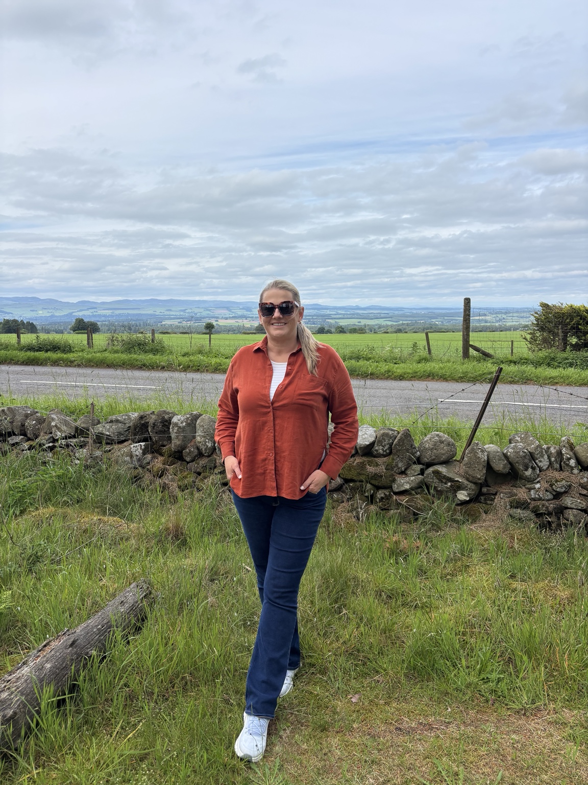 Jeni standing beside a stone wall with broad Perthshire countryside views behind her