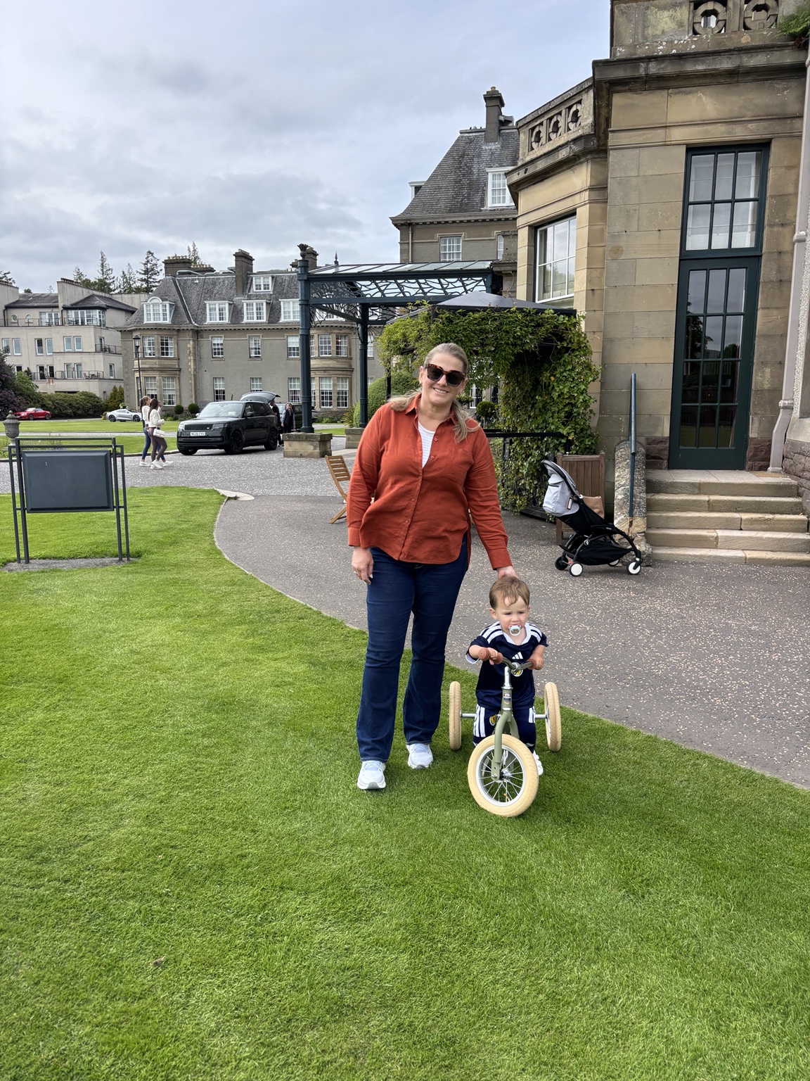 Jeni helping her toddler ride a small tricycle on the lawn at Gleneagles