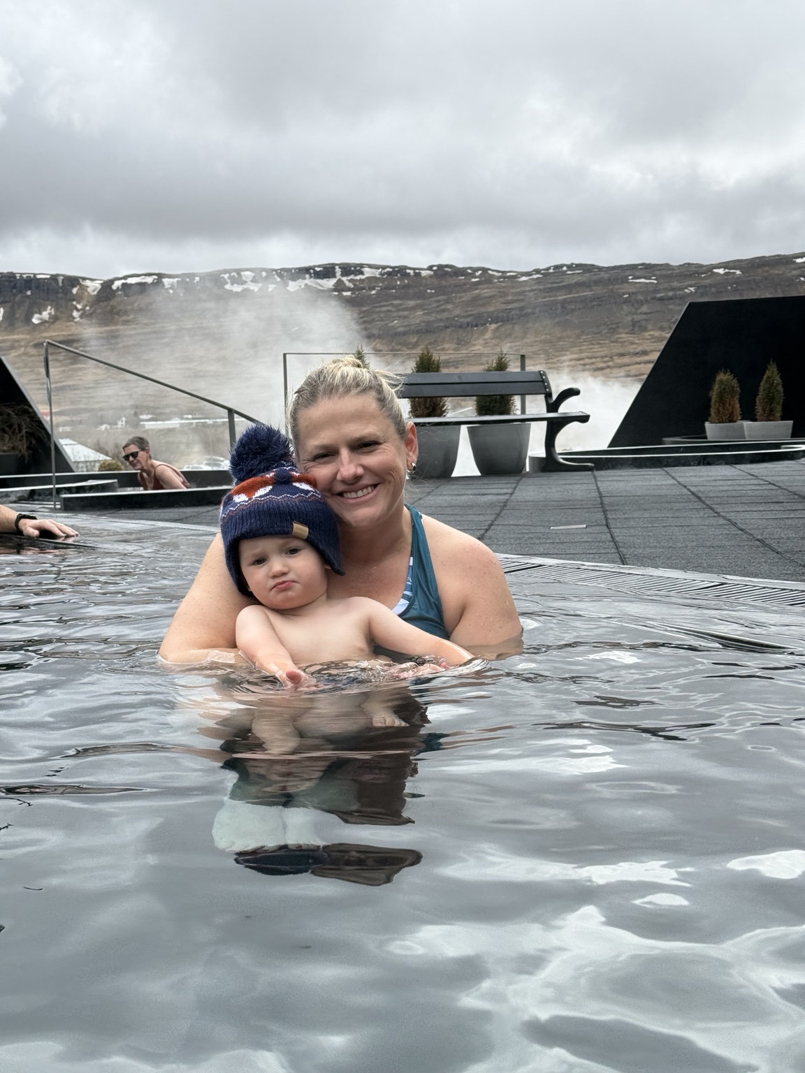 Jeni holding her toddler in a gentler-feeling pool at Krauma with steam and West Iceland hills behind them